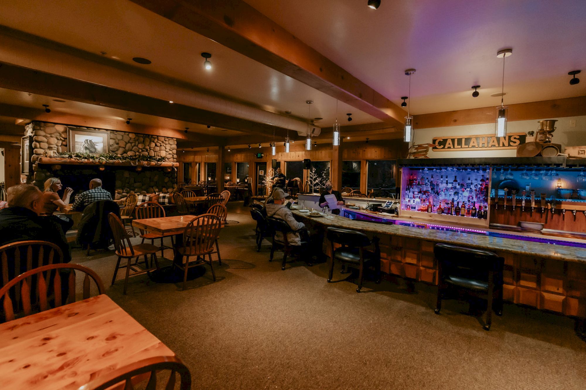 The image shows a cozy restaurant interior with a stone fireplace, wooden tables, chairs, and a bar with bottles displayed.