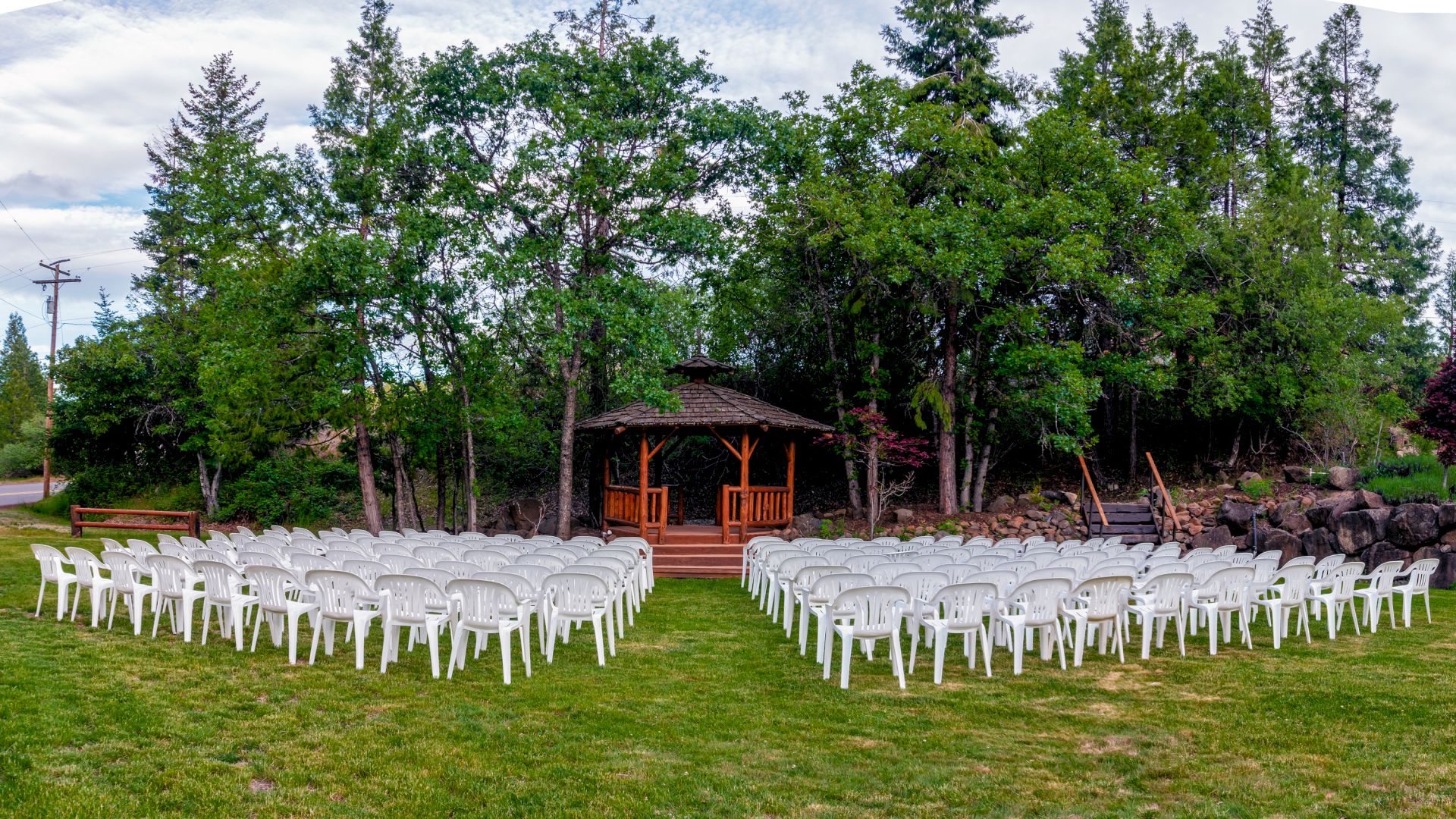 An outdoor wedding setup with rows of white chairs facing a wooden gazebo in a garden surrounded by trees and greenery.