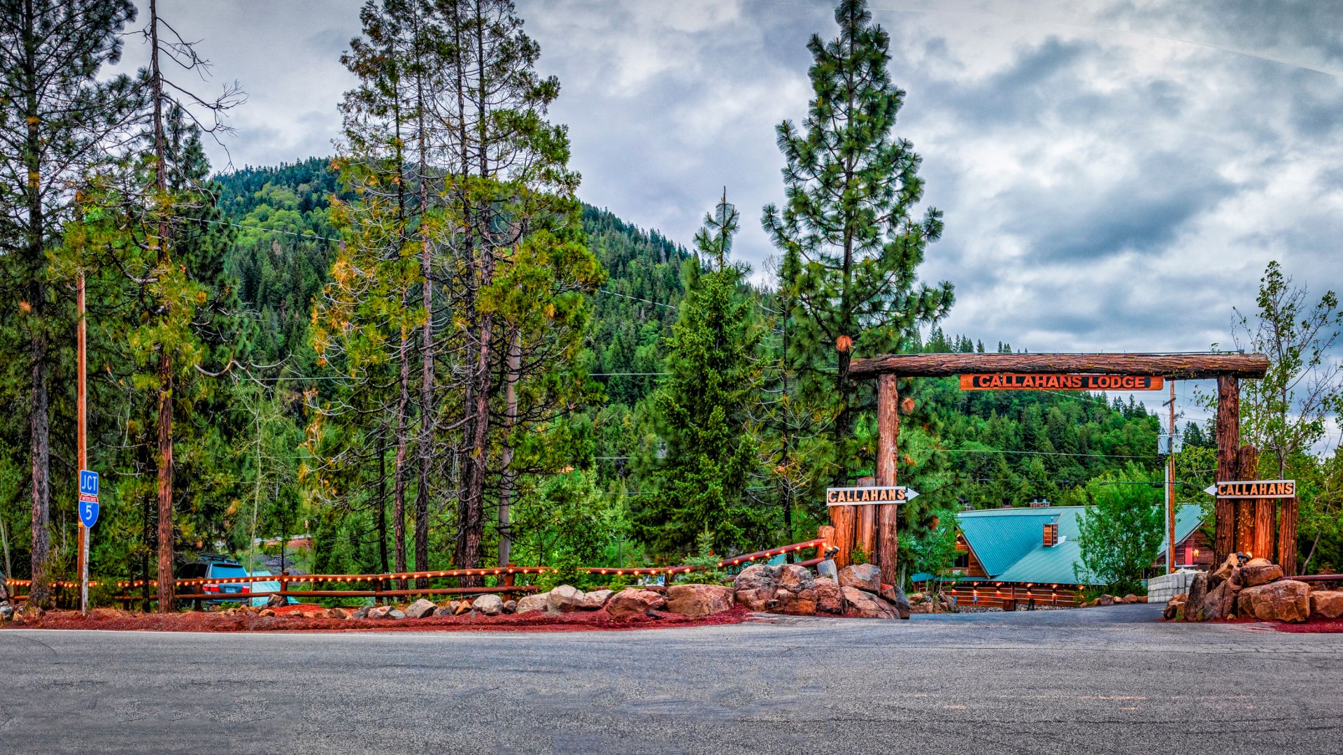 A lodge entrance amidst tall pine trees and mountains, with a wooden archway and signage, under an overcast sky.