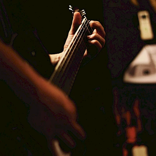 A person is playing an electric guitar in a dimly lit setting, focusing on their hand gripping the neck of the guitar and pressing the strings.