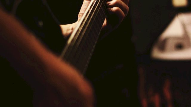 A person is playing an electric guitar in a dimly lit setting, focusing on their hand gripping the neck of the guitar and pressing the strings.