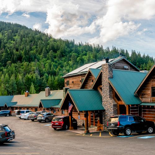 A rustic lodge with green roofs, surrounded by parked cars and lush, forested hills under a partly cloudy sky.