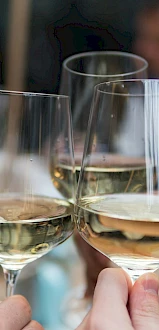 Two people clinking glasses of white wine at a table in a blurred restaurant setting, celebrating or toasting together.