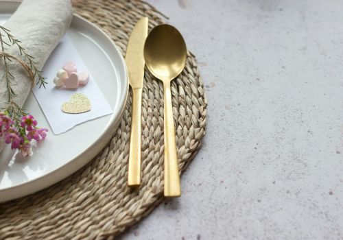 A stylish place setting with a white plate, gold cutlery, a woven placemat, and small decorative cards with hearts, plus flowers.