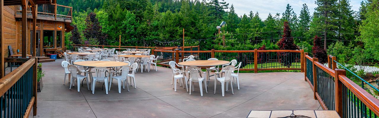 An outdoor wooden deck with several white chairs and tables, a fire pit in the foreground, and a forested hillside in the background.