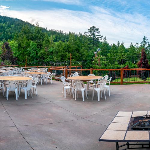 An outdoor wooden deck with several white chairs and tables, a fire pit in the foreground, and a forested hillside in the background.