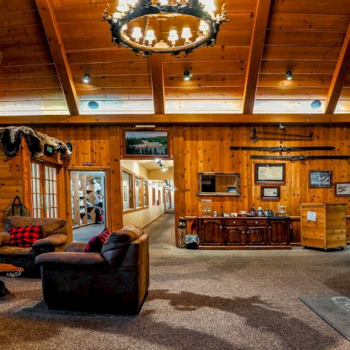 A cozy rustic lodge lobby with wooden walls, leather chairs, a chandelier, taxidermy displays, a front desk, and a warm, welcoming reception area.