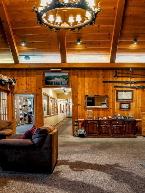 A cozy rustic lodge lobby with wooden walls, leather chairs, a chandelier, taxidermy displays, a front desk, and a warm, welcoming reception area.