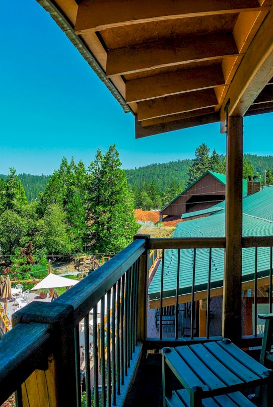 A sunny balcony view over green trees and distant hills, with wooden chairs and railing, part of a cabin or lodge.