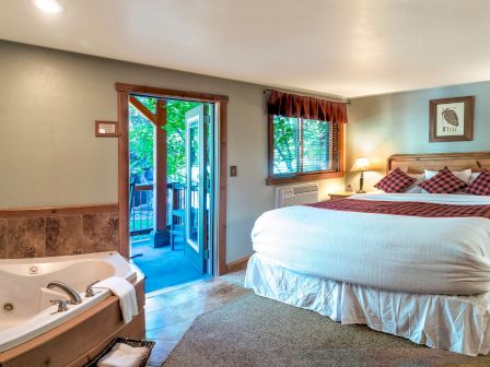 A cozy hotel room with a large bed, nightstands, lamps, a chair and desk, plus a jetted tub near an open door to a balcony.