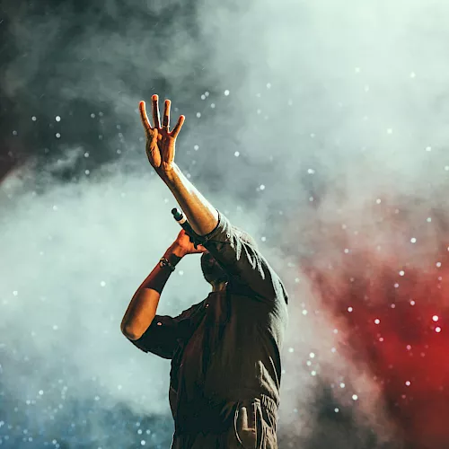 A performer on stage raises one hand amid colorful smoke and confetti, creating a dramatic live-show vibe.