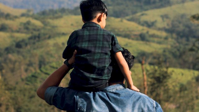 A man carries a child on his shoulders, overlooking rolling hills and distant mountains in a lush, panoramic landscape.