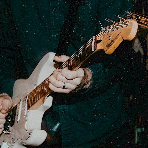 A person is playing an electric guitar or bass, gripping the fretboard as strings glow against a dark, moody background.