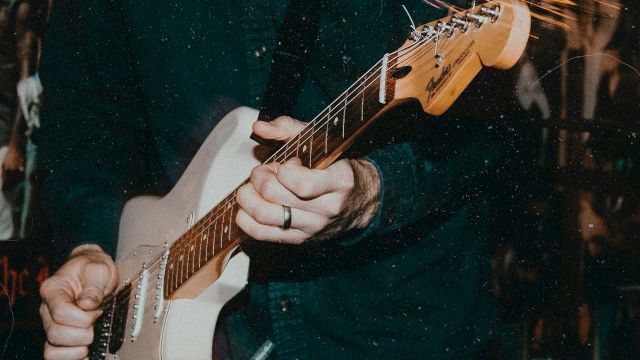 A person is playing an electric guitar or bass, gripping the fretboard as strings glow against a dark, moody background.