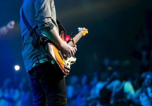 A musician performs on stage, strumming an electric guitar under blue lights.