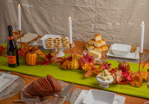 A fall-themed buffet table with lit candles, bread rolls, pastries, cheese, and drinks, surrounded by autumn leaves.