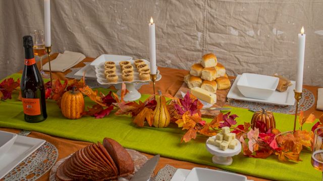 A fall-themed buffet table with lit candles, bread rolls, pastries, cheese, and drinks, surrounded by autumn leaves.