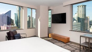 A modern hotel room with large windows showing a cityscape, a bed, a wall-mounted TV, an armchair, and a small table.