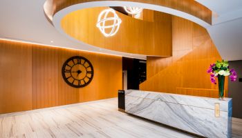The image shows a modern lobby with a marble reception desk, a large wall clock, wooden panels, and a spiral design on the ceiling with hanging lights.