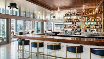 The image shows a modern indoor bar with high stools, a well-stocked bar shelf, and a view of city buildings through large windows.