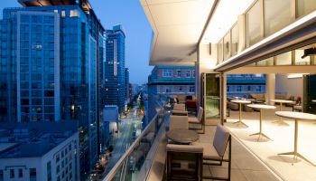 A modern cityscape during twilight, with a view from a high balcony featuring outdoor seating and tables, overlooking illuminated buildings and the street.