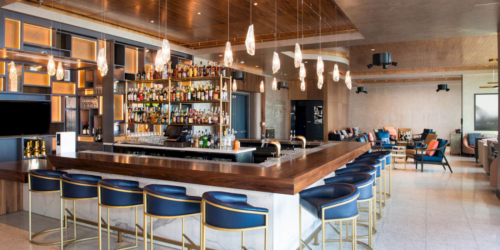 A modern bar with wooden countertops, blue barstools, hanging lights, and a well-stocked shelf of various alcohol bottles.