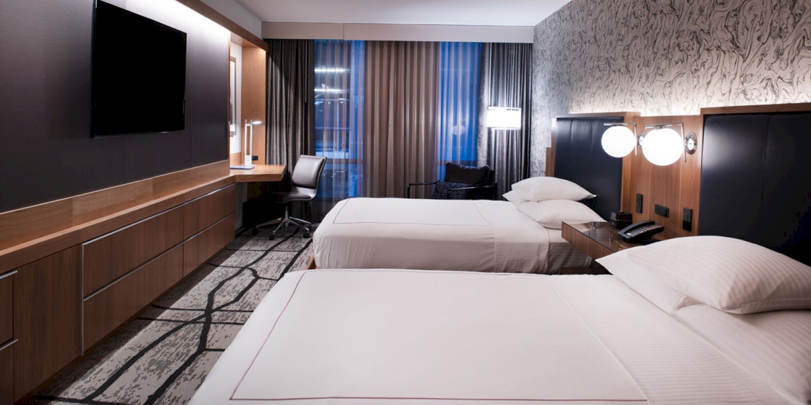 A modern hotel room with two beds, a TV, a desk and a chair by the window, and stylish décor on the walls and carpet ends the sentence.