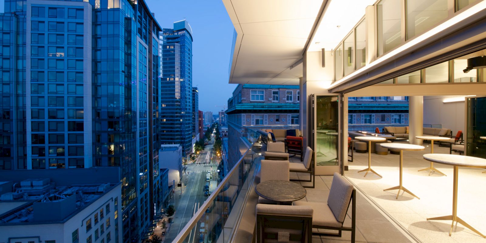 A modern cityscape view from a high-rise balcony at dusk, with seating arrangements and tables illuminated by warm indoor lighting.