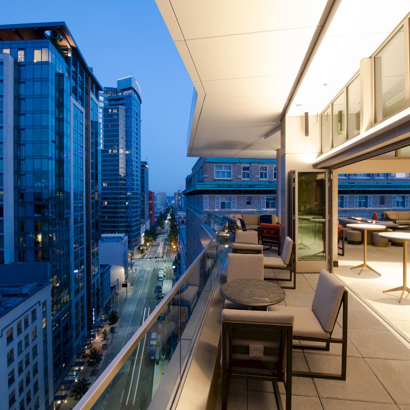 An urban balcony at twilight with modern seating, overlooking a city street and tall buildings. Indoor dining area visible inside.