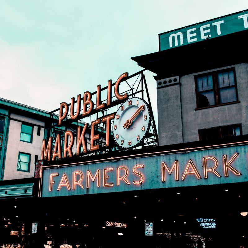 The image shows a neon lit sign for "PUBLIC MARKET" and "FARMERS MARKET" in front of several buildings, suggesting a busy marketplace.
