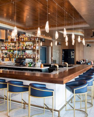 A modern bar with a wooden countertop, blue bar stools, a variety of drinks on shelves, hanging lights, and a comfortable seating area.