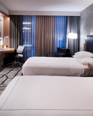 A modern hotel room with two beds, a flat-screen TV, a work desk with a chair, nightstands with lamps, and large windows with curtains.