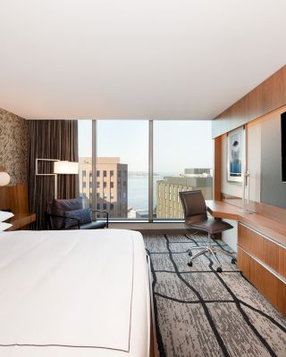 A modern hotel room with a large bed, desk, flat-screen TV, and floor-to-ceiling window providing a cityscape view outside.