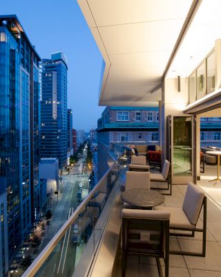 A modern rooftop terrace at dusk with seating areas overlooks a cityscape of tall buildings and street lights.