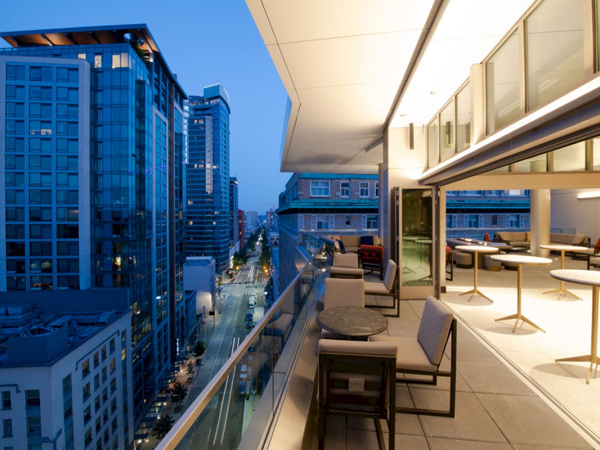 A modern balcony view of a cityscape at dusk with outdoor seating and tables, overlooking tall buildings and a street below.