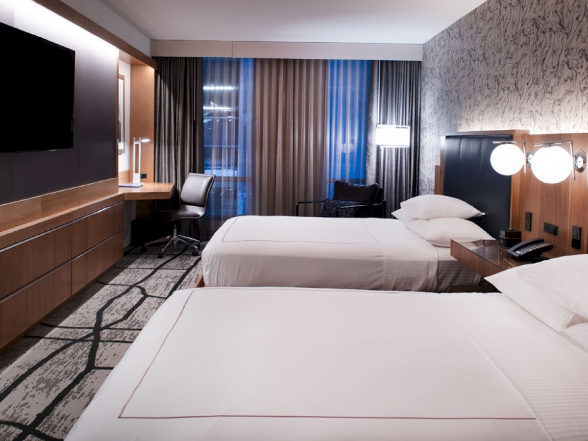 A modern hotel room with two double beds, a flat-screen TV, a work desk with a chair, bedside lamps, and patterned carpet and wallpaper.