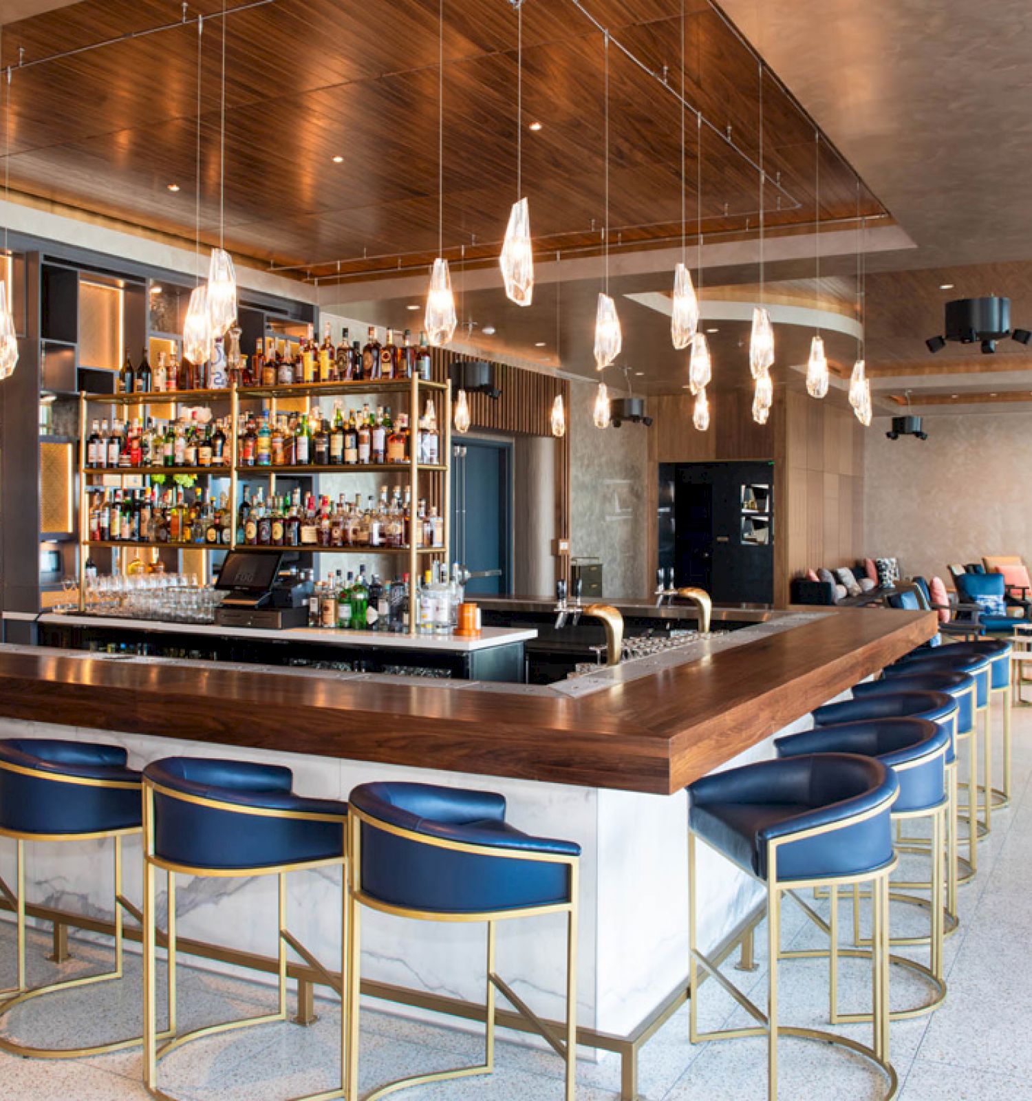 A stylish bar with modern lighting, blue chairs, a wide selection of bottles, and a sleek design, including a lounge area in the background.