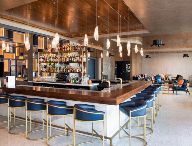 The image shows a modern bar with stylish blue chairs, a well-stocked back bar, hanging lights, and a lounge area in the background.