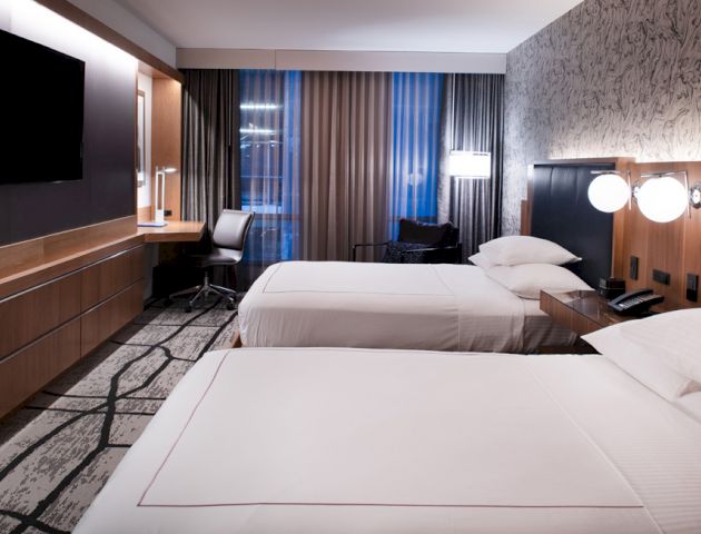 A modern hotel room features two neatly made beds, a flat-screen TV, a desk with a chair, wall lamps, and a stylish decor with patterned wallpaper.