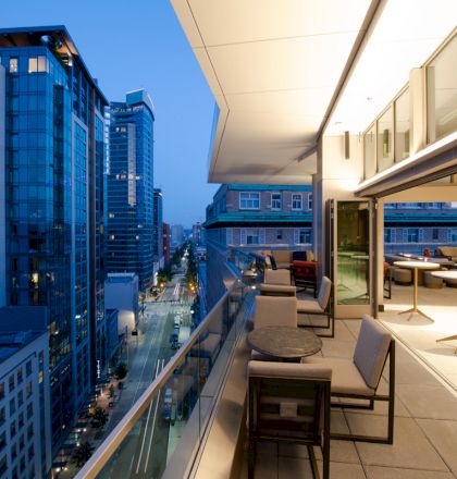 A modern balcony overlooks a cityscape at dusk, featuring outdoor seating and tables under a well-lit covered area, with high-rise buildings in view.