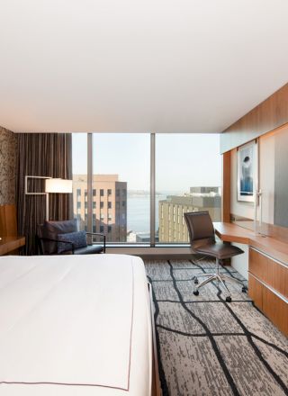 A modern hotel room with a large bed, wall-mounted TV, desk, chair, and floor-to-ceiling windows offering a city view.