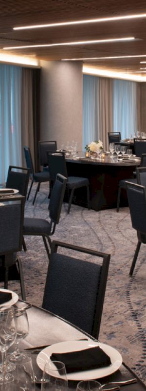 The image shows a modern banquet hall set for an event, with round tables and chairs, neatly arranged place settings, and soft lighting.
