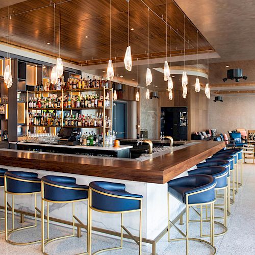 This image shows a modern bar with a central counter surrounded by blue and gold stools, a variety of drinks on shelves, and contemporary decor.