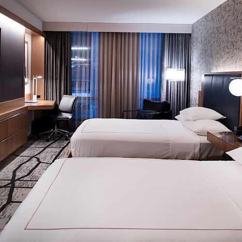 The image shows a modern hotel room with two neatly made beds, a wall-mounted TV, a desk with a chair, and large curtains over windows.