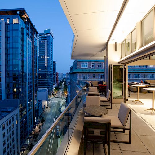 A modern rooftop patio at dusk overlooks a cityscape of tall buildings and busy streets, featuring outdoor seating and an inviting ambiance.