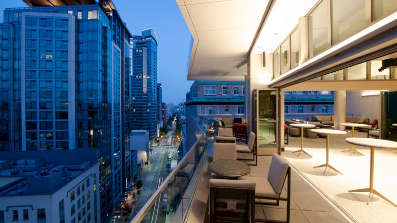 A modern cityscape at dusk with tall buildings, viewed from an open balcony with tables, chairs, and lights, creating an inviting atmosphere.