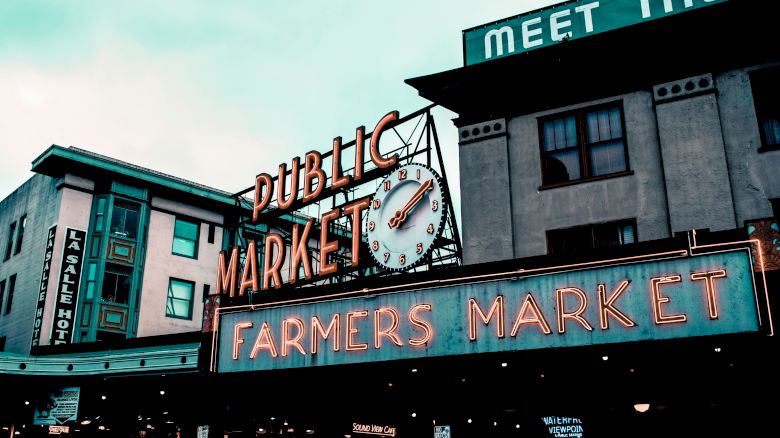 The image depicts the iconic entrance sign of a Public Market with a clock and "FARMERS MARKET" written below in neon lights.