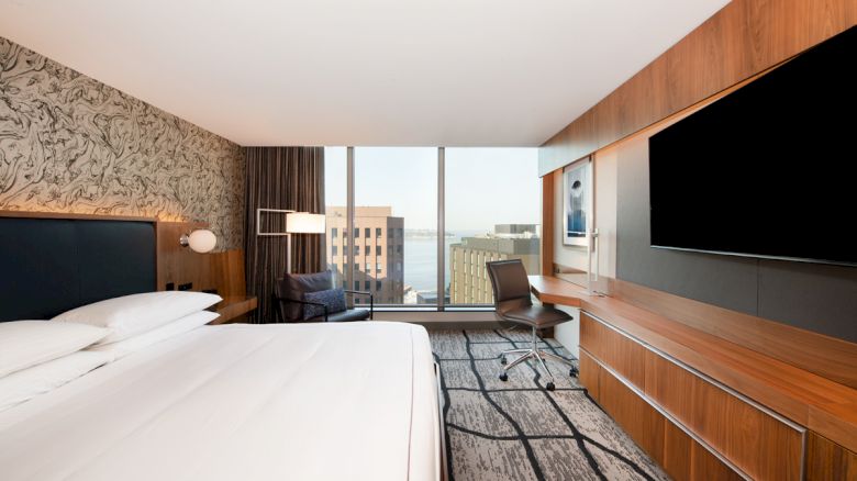 A modern hotel room with a large bed, a wall-mounted TV, a desk, a chair, an artistic wall design, and a window with a city view.
