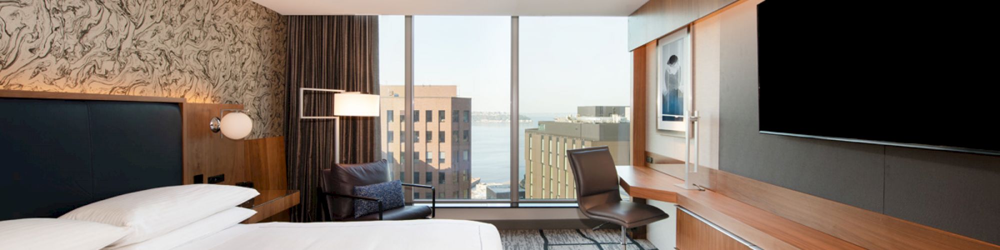 A modern hotel room with a large bed, wall-mounted TV, desk, and chair, featuring a large window with a cityscape view in the background.