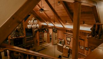 A cozy wooden cabin interior with high ceilings, stone fireplace, chandelier, and rustic decor, viewed from a lofted perspective.